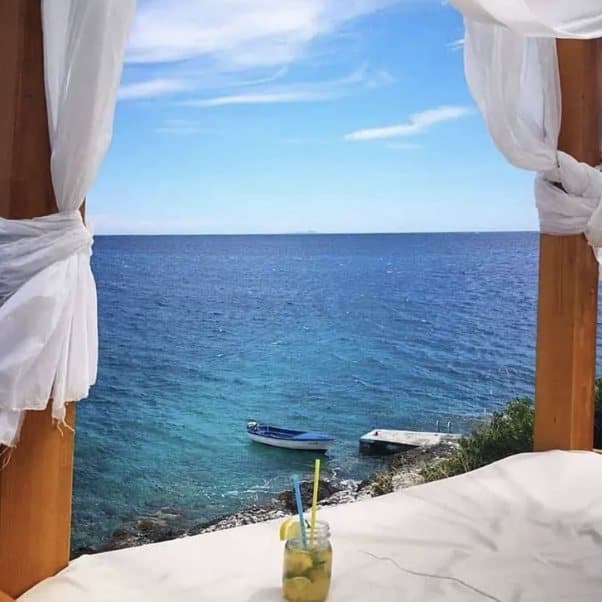 View from a canopy bed at Pacha Mama — white curtains, lemonade on the cushion, a boat and the pontoon in the turquoise bay below